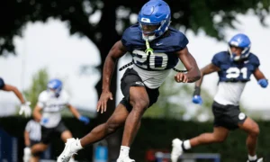 Football players in blue helmets sprint across a practice field, central player in navy jersey (#10) leading the charge.