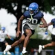 Football players in blue helmets sprint across a practice field, central player in navy jersey (#10) leading the charge.