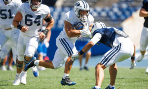 Football players in white uniforms chase a ball carrier during a daytime game; BYU logo visible on the helmet.