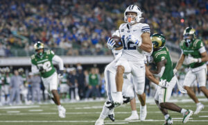 BYU football player in white uniform carries the ball, pursued by green-clad defenders on a crowded stadium field.