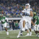 BYU football player in white uniform carries the ball, pursued by green-clad defenders on a crowded stadium field.
