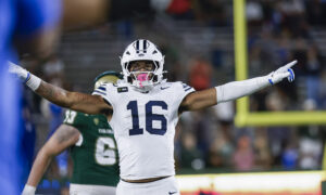 BYU football player in white jersey number 16 raises arms in celebration near end zone, helmet on, stadium crowd in background.