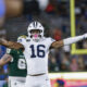 BYU football player in white jersey number 16 raises arms in celebration near end zone, helmet on, stadium crowd in background.