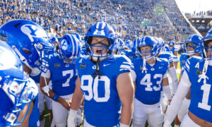 BYU football players in blue uniforms form a huddle on the field with a packed stadium in the background.