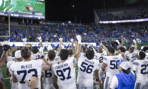 Huddle of football players in white jerseys on the field while a packed blue-clad stadium cheers at night, under bright lights.
