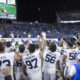 Huddle of football players in white jerseys on the field while a packed blue-clad stadium cheers at night, under bright lights.