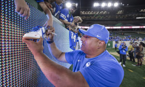 Man in a blue shirt signs a white Nike sneaker against an LED barrier as fans lean over to watch at a stadium.