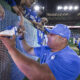 Man in a blue shirt signs a white Nike sneaker against an LED barrier as fans lean over to watch at a stadium.