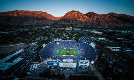 Aerial view of a full football stadium with BYU markings, set against rugged mountains at dusk in Utah.
