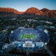 Aerial view of a full football stadium with BYU markings, set against rugged mountains at dusk in Utah.