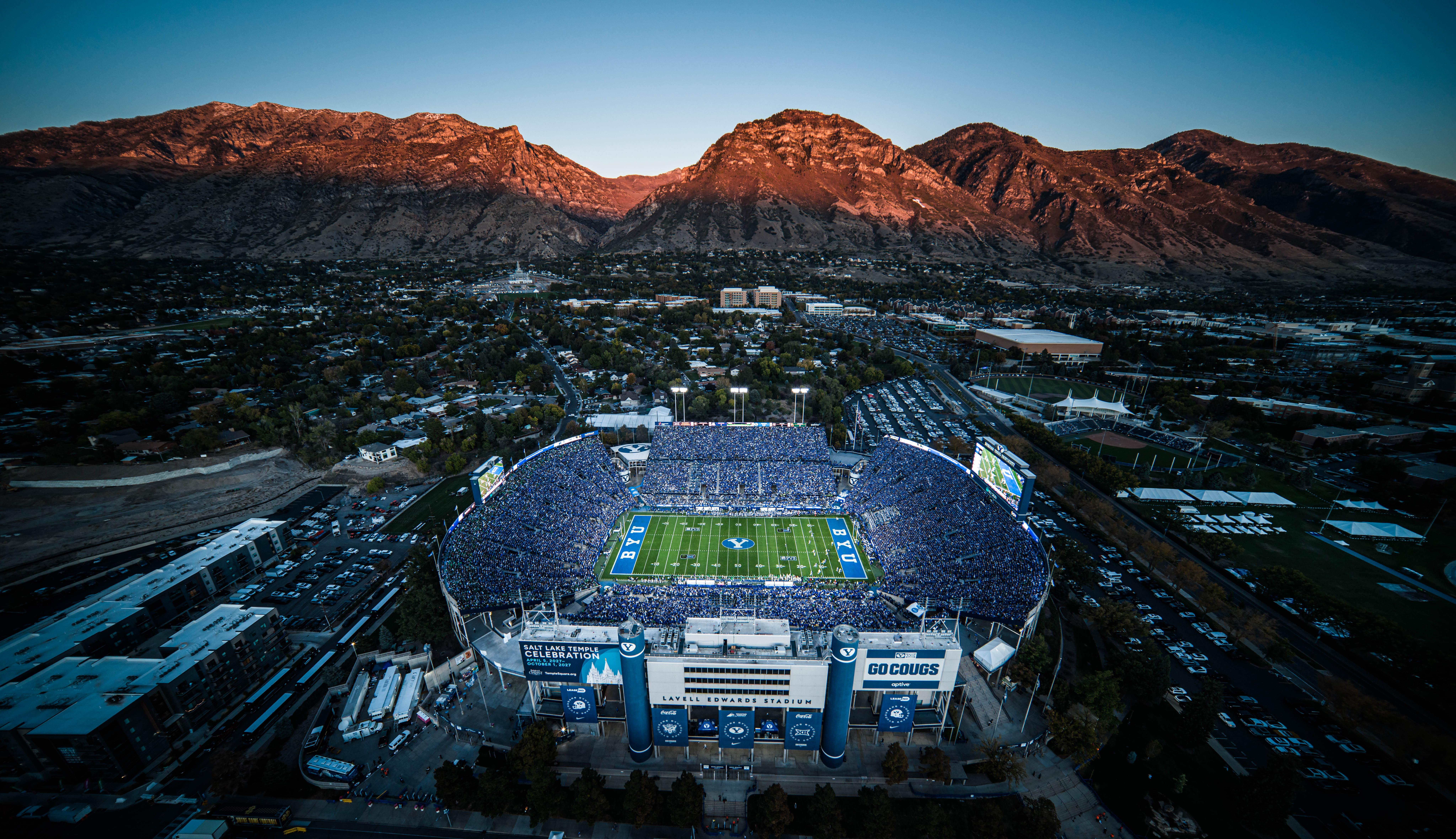Aerial view of a full football stadium with BYU markings, set against rugged mountains at dusk in Utah.