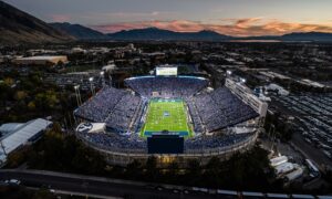 Aerial view of a packed football stadium at dusk, with bright field lights and a surrounding cityscape and distant mountains under a sunset sky.