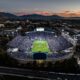 Aerial view of a packed football stadium at dusk, with bright field lights and a surrounding cityscape and distant mountains under a sunset sky.