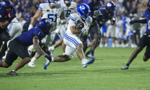 A football running back in a white and blue uniform carries the ball as defenders in blue pursue on a grassy field during a game.