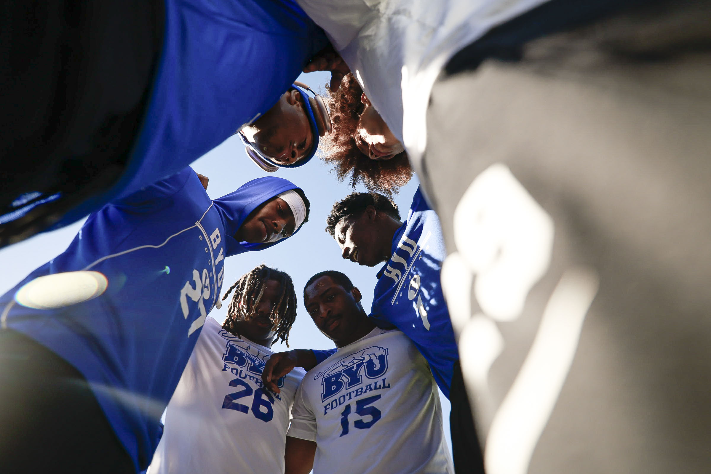 Group of football players in blue and white jerseys forming a low huddle, heads together, viewed from below]