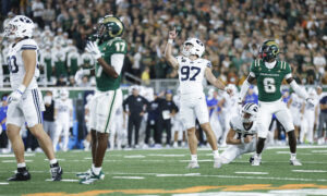 Football players in white and green uniforms on a crowded field, with a player raising his arm in celebration as the crowd cheers in the background.
