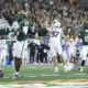 Football players in white and green uniforms on a crowded field, with a player raising his arm in celebration as the crowd cheers in the background.