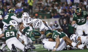 Group of football players in green and white pile up on the field during a game, with spectators blurred in the background.