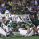 Group of football players in green and white pile up on the field during a game, with spectators blurred in the background.