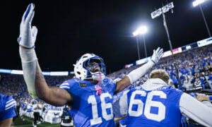 American football players in blue uniforms celebrate with raised arms on the sideline under bright stadium lights, crowd cheering in the background at night.