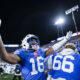 American football players in blue uniforms celebrate with raised arms on the sideline under bright stadium lights, crowd cheering in the background at night.