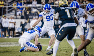 BYU football players tackle on a green field during a game, with a blue-helmeted player in white jersey (#97) in focus and a defender in dark uniform nearby; stadium crowd in the background