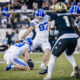 BYU football players tackle on a green field during a game, with a blue-helmeted player in white jersey (#97) in focus and a defender in dark uniform nearby; stadium crowd in the background