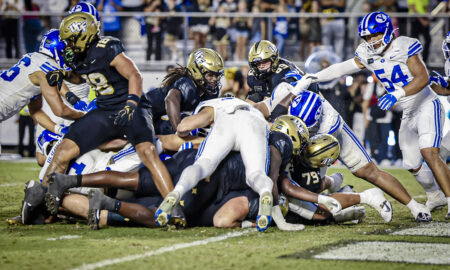 Football players collide in a pile on the field during a game, gold helmets and blue-and-white uniforms visible in a chaotic scrum.