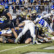 Football players collide in a pile on the field during a game, gold helmets and blue-and-white uniforms visible in a chaotic scrum.