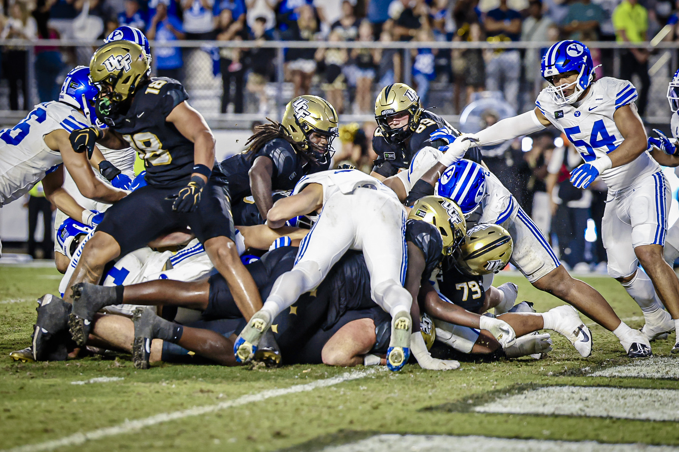Football players collide in a pile on the field during a game, gold helmets and blue-and-white uniforms visible in a chaotic scrum.