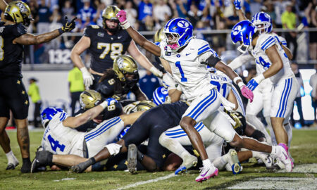 Action shot of players colliding on a football field, blue-and-white uniforms in the foreground and a crowded stadium in the background.