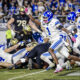 Action shot of players colliding on a football field, blue-and-white uniforms in the foreground and a crowded stadium in the background.