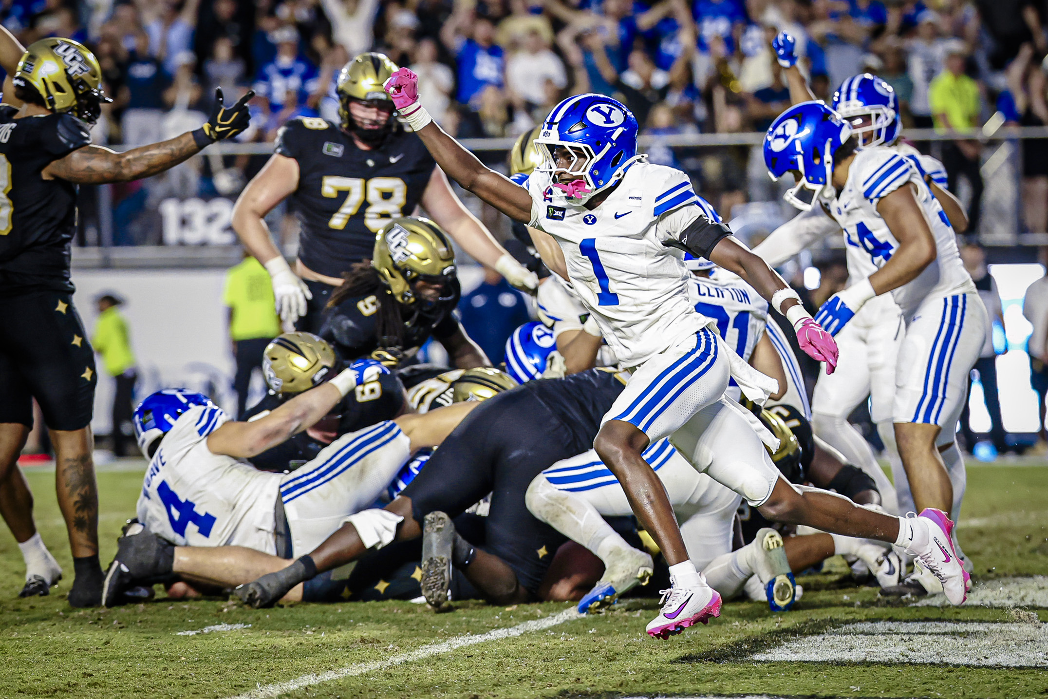 Action shot of players colliding on a football field, blue-and-white uniforms in the foreground and a crowded stadium in the background.