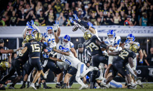 Group of football players in white, blue, and black uniforms collide on the field as fans cheer in the stands.