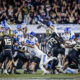 Group of football players in white, blue, and black uniforms collide on the field as fans cheer in the stands.