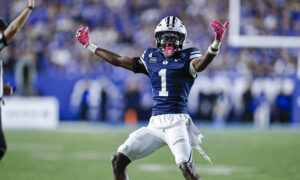 BYU football player in blue jersey (number 1) with white helmet raises arms in celebration on a green field.