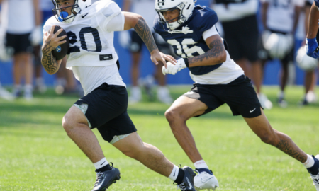 Two football players sprint on a sunny practice field; ball carrier in white jersey #80 leads as a defender in blue #35 pursues close behind.