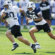Two football players sprint on a sunny practice field; ball carrier in white jersey #80 leads as a defender in blue #35 pursues close behind.