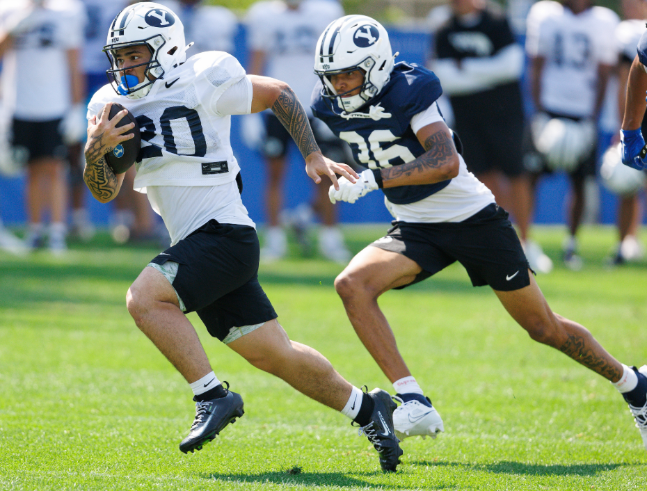 Two football players sprint on a sunny practice field; ball carrier in white jersey #80 leads as a defender in blue #35 pursues close behind.