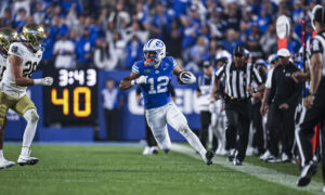 BYU football player #12 runs with the ball while Notre Dame defenders pursue; crowded stadium in background.