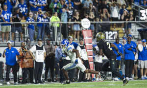 Two football players sprint toward the end zone on a green field, with a cheering crowd in blue jerseys in the stands behind them.