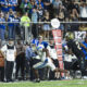 Two football players sprint toward the end zone on a green field, with a cheering crowd in blue jerseys in the stands behind them.
