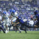 BYU and TCU players chase a ball carrier during a crowded football game on a green field, with defenders in purple pursuing from the right and white-uniformed players on the left.