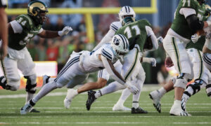 College football players in green and white uniforms collide during a tackle on a green turf field.