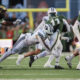 College football players in green and white uniforms collide during a tackle on a green turf field.