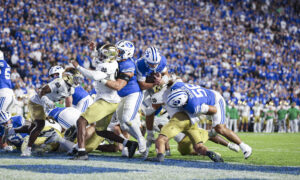 College football players from BYU and Notre Dame clash for the ball as fans fill the blue stadium seating behind them.