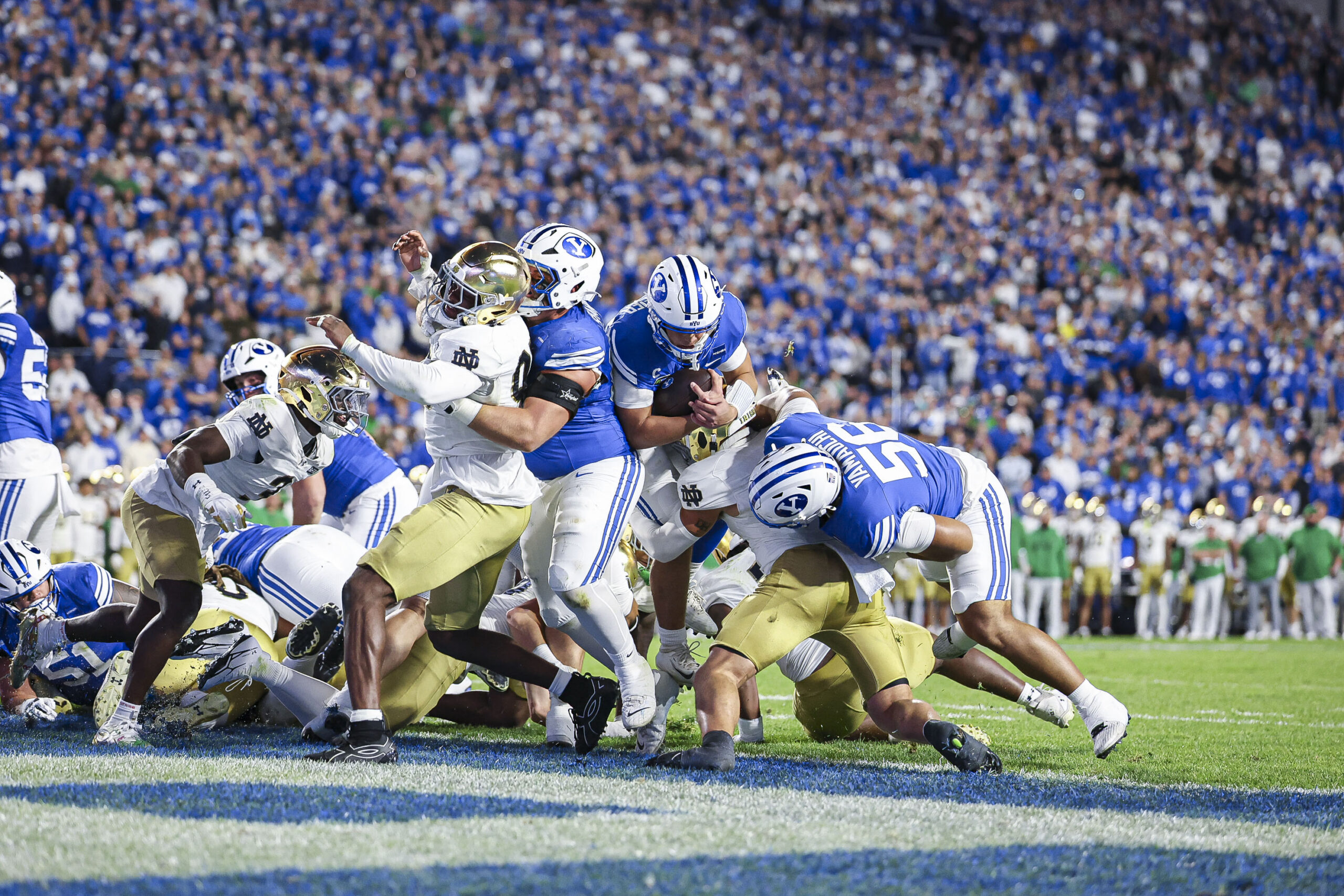 College football players from BYU and Notre Dame clash for the ball as fans fill the blue stadium seating behind them.