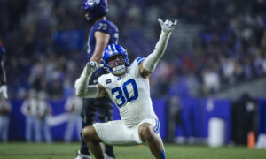 BYU football player in blue and white uniform celebrating with arms raised on the field, crowd blurred in background