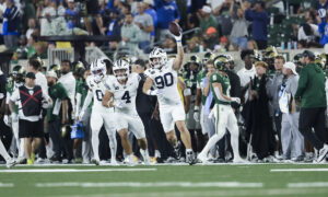 White-uniform football players celebrate on the field as a teammate raises a ball, with fans in the stands behind them.
