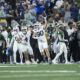 White-uniform football players celebrate on the field as a teammate raises a ball, with fans in the stands behind them.